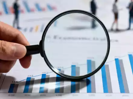 Eine Hand hält eine Lupe über einem Balkendiagramm, das Wirtschaftsdaten darstellt, mit unscharfen Diagrammen im Hintergrund. - Foto: über boerse-global.de