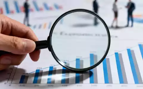Eine Hand hält eine Lupe über einem Balkendiagramm, das Wirtschaftsdaten darstellt, mit unscharfen Diagrammen im Hintergrund. - Foto: über boerse-global.de