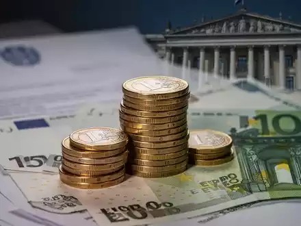 Ein Nahaufnahme-Bild von gestapelten Euro-Münzen und Banknoten, mit unscharfem Hintergrund von Dokumenten und einem Regierungsgebäude. - Foto: über boerse-global.de