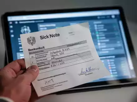 Eine Hand hält eine Krankmeldung aus Papier. Im unscharfen Hintergrund ist ein Tablet mit medizinischen Daten zu sehen, das den digitalen Wandel symbolisiert. - Foto: über boerse-global.de