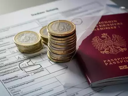 Ein Stapel Euro-Münzen und ein niederländischer Reisepass, teilweise verdeckt von deutschen Steuerformularen, symbolisieren grenzüberschreitende Finanzen. - Foto: über boerse-global.de