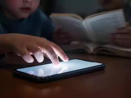 Kinderhand greift nach leuchtendem Smartphone, während andere Hand ein unscharfes Buch hält, symbolisiert digitale Ablenkung. - Foto: über boerse-global.de
