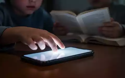 Kinderhand greift nach leuchtendem Smartphone, während andere Hand ein unscharfes Buch hält, symbolisiert digitale Ablenkung. - Foto: über boerse-global.de