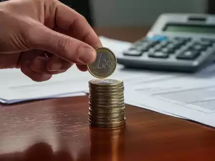 Eine Hand legt eine Euro-Münze auf einen Stapel Münzen, im Hintergrund verschwommen ein Taschenrechner und Dokumente. - Foto: über boerse-global.de
