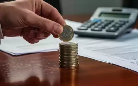 Eine Hand legt eine Euro-Münze auf einen Stapel Münzen, im Hintergrund verschwommen ein Taschenrechner und Dokumente. - Foto: über boerse-global.de