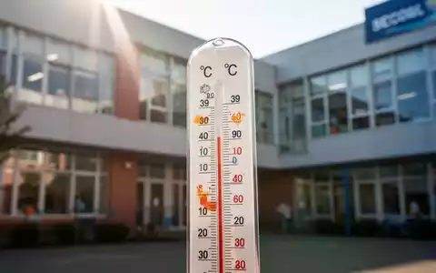 Ein Thermometer zeigt hohe Temperaturen vor der unscharfen Fassade eines modernen Schulgebäudes. - Foto: über boerse-global.de