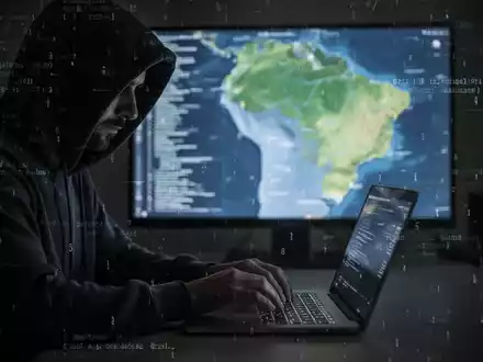 A shadowy figure in a hoodie types on a laptop, with a blurred map of Brazil and digital code in the background, symbolizing a cyberattack. - Foto: über boerse-global.de