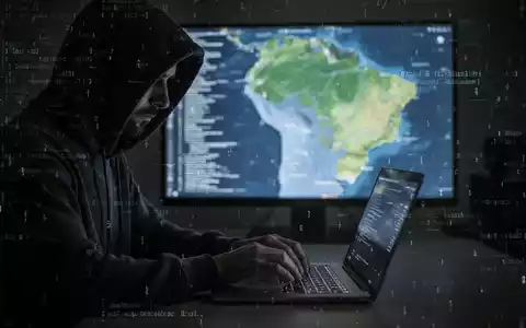 A shadowy figure in a hoodie types on a laptop, with a blurred map of Brazil and digital code in the background, symbolizing a cyberattack. - Foto: über boerse-global.de