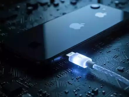 A close-up of an Apple iPhone connected to a circuit board by a glowing USB cable, symbolizing a hardware exploit. - Foto: über boerse-global.de