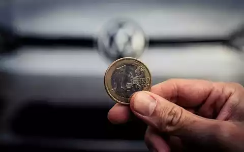 Nahaufnahme einer Hand, die eine Euromünze hält, mit einem unscharfen Volkswagen-Emblem im Hintergrund. - Foto: über boerse-global.de