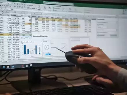 A close-up of a hand using a computer mouse on a desk, with a monitor in the background displaying an Excel spreadsheet with data and charts. - Foto: über boerse-global.de
