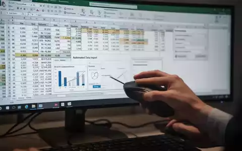 A close-up of a hand using a computer mouse on a desk, with a monitor in the background displaying an Excel spreadsheet with data and charts. - Foto: über boerse-global.de