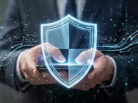 A glowing digital shield icon overlaid on a blurred image of hands using a smartphone, with data streams. - Foto: über boerse-global.de