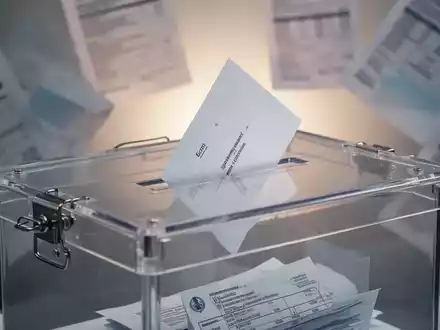 Eine transparente Wahlurne mit einem einzelnen Stimmzettel vor einem verschwommenen Hintergrund von Finanzdokumenten. - Foto: über boerse-global.de