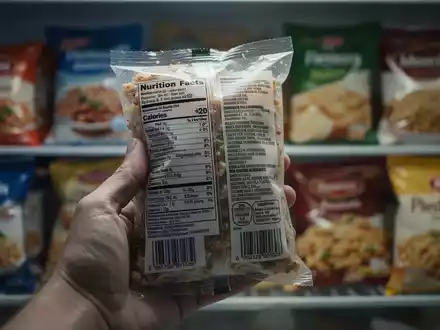 Nahaufnahme einer Hand, die eine Lebensmittelverpackung hält, mit verschwommenem Hintergrund von verarbeiteten Lebensmitteln. - Foto: über boerse-global.de