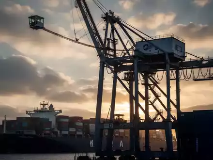 Ein riesiger Hafen-Containerkran mit einer kleinen Kabine in großer Höhe, bei Sonnenaufgang oder -untergang. - Foto: über boerse-global.de