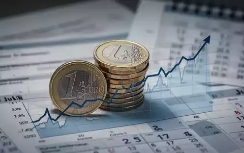 Ein Stapel deutscher Euro-Münzen vor einem Finanzdiagramm, das einen Aufwärtstrend zeigt, mit verschwommenen Dokumenten und einem Kalender im Hintergrund. - Foto: über boerse-global.de