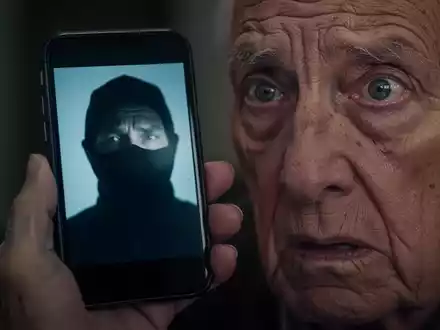 Eine ältere Person hält ein Smartphone, auf dessen Bildschirm das unscharfe Gesicht eines Betrügers zu sehen ist, das sich im besorgten Auge spiegelt. - Foto: über boerse-global.de