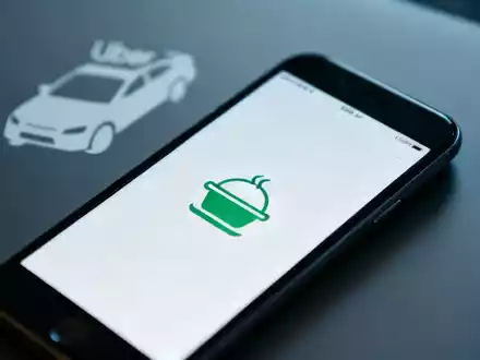 Smartphone mit einer Essensliefer-App, im Hintergrund verschwommen ein Uber-Auto-Symbol, das eine Beteiligung andeutet. - Foto: über boerse-global.de
