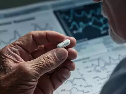Eine ältere Hand hält eine Pille, im unscharfen Hintergrund sind medizinische Diagramme und Daten erkennbar. - Foto: über boerse-global.de
