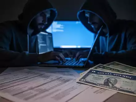 A shadowy figure typing on a laptop, with blurred financial documents and a Social Security card in the foreground. - Foto: über boerse-global.de