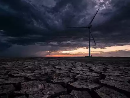 Eine einzelne Windturbine vor einem stürmischen Himmel, der Boden ist rissig und trocken. Symbolisiert Druck nach starkem Lauf. - Foto: über boerse-global.de