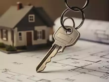 Ein Schlüsselbund mit einem einzelnen Schlüssel, der vor einem unscharfen Hintergrund aus Bauplänen oder einem kleinen Modellhaus liegt. - Foto: über boerse-global.de