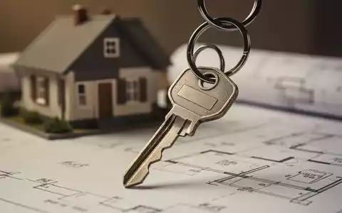 Ein Schlüsselbund mit einem einzelnen Schlüssel, der vor einem unscharfen Hintergrund aus Bauplänen oder einem kleinen Modellhaus liegt. - Foto: über boerse-global.de