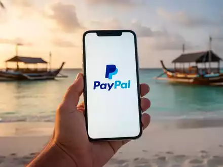 A hand holding a smartphone showing the PayPal logo, with a blurred background of a Maldivian beach and boats. - Foto: über boerse-global.de