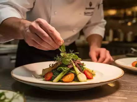 Ein Koch garniert ein kunstvoll angerichtetes vegetarisches Gericht in einer professionellen Küche mit sanftem Licht. - Foto: über boerse-global.de