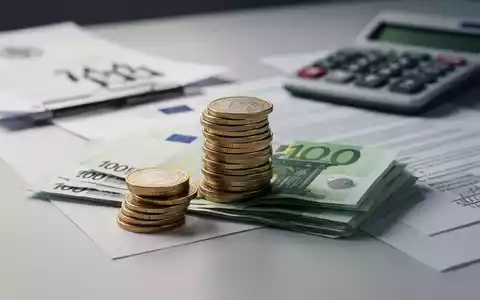 Ein Stapel Euro-Münzen und -Scheine auf einem Schreibtisch, mit einem Taschenrechner und Dokumenten im unscharfen Hintergrund. - Foto: über boerse-global.de