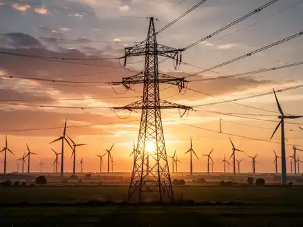 Hochspannungsmast im Sonnenuntergang mit Windturbinen im Hintergrund, symbolisiert RWEs Beteiligung an Amprion. - Foto: über boerse-global.de