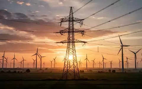 Hochspannungsmast im Sonnenuntergang mit Windturbinen im Hintergrund, symbolisiert RWEs Beteiligung an Amprion. - Foto: über boerse-global.de