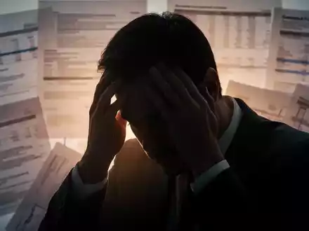 Eine Person sitzt mit dem Kopf in den Händen, vor einem unscharfen Hintergrund aus Finanzdokumenten, der psychische Belastung darstellt. - Foto: über boerse-global.de