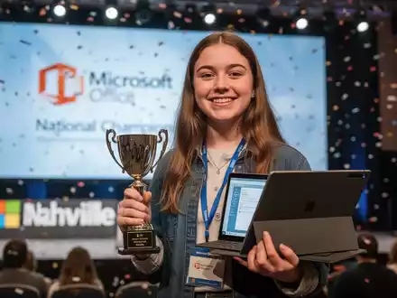 A young woman, Rebecca Sutherlin, holds a trophy and a Surface Pro laptop, smiling, on a competition stage. - Foto: über boerse-global.de