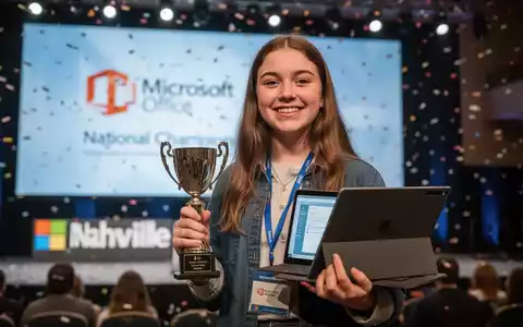 A young woman, Rebecca Sutherlin, holds a trophy and a Surface Pro laptop, smiling, on a competition stage. - Foto: über boerse-global.de