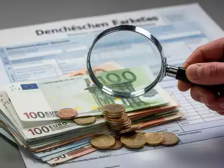 Eine Hand hält eine Lupe über Euro-Banknoten und Münzen; im Hintergrund ein unscharfes deutsches Steuerformular. - Foto: über boerse-global.de