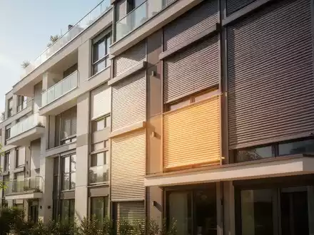 Eine moderne Wohnhausfassade mit teilweise heruntergelassenen Rollläden an den Fenstern, die Licht durchlassen. - Foto: über boerse-global.de