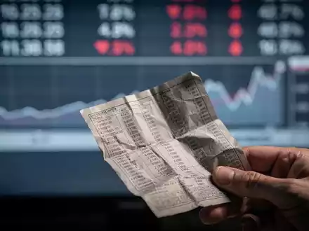 Eine Hand hält ein zerknittertes Finanzdokument vor einem unscharfen Börsenticker, der einen leichten Rückgang anzeigt. - Foto: über boerse-global.de