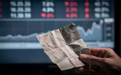 Eine Hand hält ein zerknittertes Finanzdokument vor einem unscharfen Börsenticker, der einen leichten Rückgang anzeigt. - Foto: über boerse-global.de