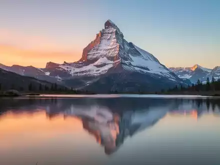 Ein majestätischer, silberner Berggipfel spiegelt sich bei Sonnenaufgang in einem ruhigen See wider. - Foto: über boerse-global.de