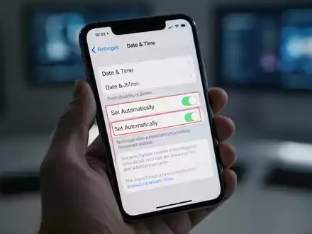 A hand holding an iPhone displaying the 'Date & Time' settings screen, with the 'Set Automatically' toggle highlighted. - Foto: über boerse-global.de