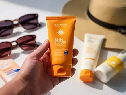 Eine Hand hält eine Tube Sonnencreme, im Hintergrund sind unscharf weitere Sonnenschutzprodukte wie Sonnenbrillen und ein Sonnenhut zu sehen. - Foto: über boerse-global.de