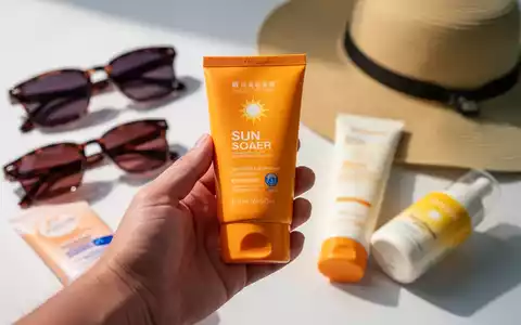 Eine Hand hält eine Tube Sonnencreme, im Hintergrund sind unscharf weitere Sonnenschutzprodukte wie Sonnenbrillen und ein Sonnenhut zu sehen. - Foto: über boerse-global.de