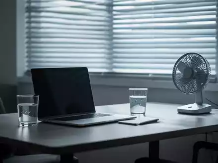 Ein modernes Homeoffice mit Laptop, Wasserglas und Ventilator vor einem Fenster mit heruntergelassenen Jalousien, das Hitzeschutz symbolisiert. - Foto: über boerse-global.de