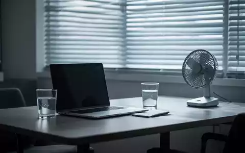 Ein modernes Homeoffice mit Laptop, Wasserglas und Ventilator vor einem Fenster mit heruntergelassenen Jalousien, das Hitzeschutz symbolisiert. - Foto: über boerse-global.de