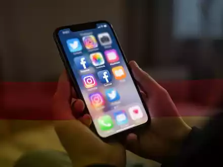 Eine junge Person hält ein Smartphone mit verschwommenen Social-Media-Symbolen auf dem Bildschirm, überlagert von den deutschen Farben. - Foto: über boerse-global.de