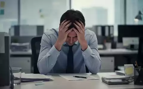 Eine Person sitzt am Schreibtisch, den Kopf in den Händen, umgeben von verschwommenen Büroeinrichtungen, die Erschöpfung symbolisieren. - Foto: über boerse-global.de