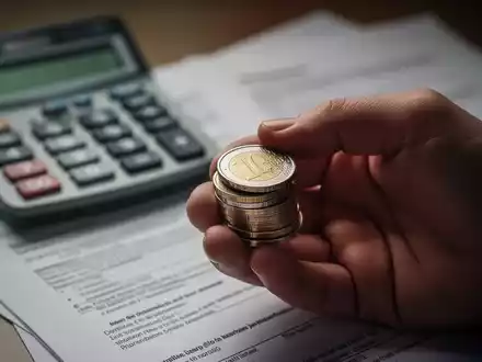 Nahaufnahme einer Hand, die Euro-Münzen hält, mit unscharfem Hintergrund von Taschenrechner und Dokumenten, die finanzielle Anpassungen symbolisieren. - Foto: über boerse-global.de
