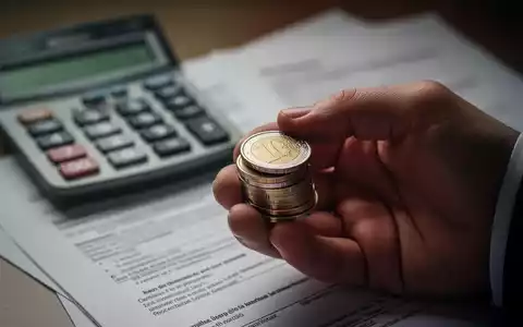 Nahaufnahme einer Hand, die Euro-Münzen hält, mit unscharfem Hintergrund von Taschenrechner und Dokumenten, die finanzielle Anpassungen symbolisieren. - Foto: über boerse-global.de
